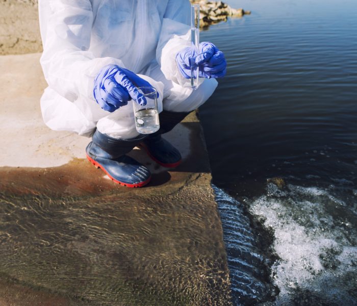Unrecognizable ecologist standing where sewage waste water meets the river and taking samples to determine level of contamination and pollution.